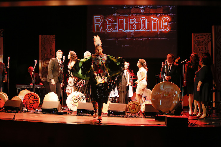 A woman in indigenous garb standing on a stage surrounded by hand-drums painted with indigenous symbols and motifs